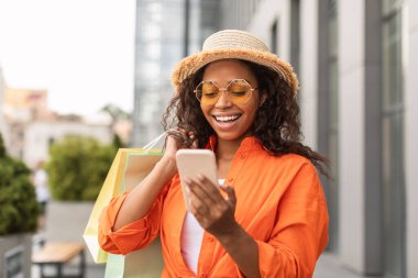 Bin yıllık Afro-Amerikan bayanı rahat, şapkalı ve gözlüklü ve akıllı telefonuyla akıllı telefondan yazı yazan mutlu yıllar. Büyük indirim, mağazadan reklam ve teklif, online alışveriş uygulaması.