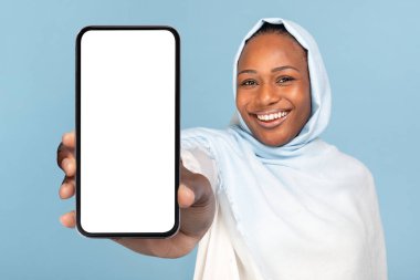 Beyaz ekranlı akıllı telefon görüntüsü. Afrikalı Amerikalı tesettürlü Müslüman kadın mavi stüdyonun arka planında duruyor. Seçici odaklanma, yakın plan.