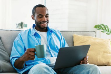 Laptop kullanan, elinde fincan tutan, sabah kahvesini içen, kanepede oturup haber okuyan gülümseyen siyah adamın portresi. Pozitif Afro-Amerikalı adam video izliyor, bilgisayar ekranına bakıyor.