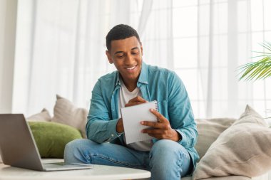 Kot pantolonlu, gülümseyen, yakışıklı siyah erkek öğrenci notlar alıyor, parlak oturma odasının içinde dizüstü bilgisayarla çalışıyor. İş, serbest çalışma, öğrenim ve covid-19 salgını sırasında evde yeni normaller için aygıt