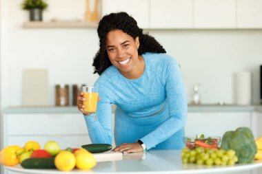 Sağlıklı beslenme konsepti. Spor giyinen, meyve suyu içen, kilo vermek için yemek pişiren, mutfakta durup gülümseyen pozitif siyah bir kadın. Diyet ve sağlık ve zayıflama için beslenme