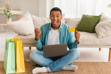 Dizüstü bilgisayarı olan mutlu genç siyahi bir adam, bir sürü alışveriş torbası büyük bir satışa seviniyor, oturma odasının içinde yerde oturuyor. Harika online alışveriş, nakit para, kazanma, alışveriş, teslimat.