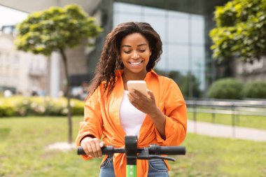 Milenyum Afrikalı Amerikalı kadın elektrikli scooterla sosyal medyada akıllı telefondan sohbet ediyor. Çevre taşımacılığı, parkta yürüyüş, yeni blog ve uygulama, kentsel hizmet