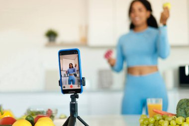 Yemek ve spor vlog 'u. Sportif siyahi kadın akıllı telefondan video çekiyor. Donut ve elmayı gösteriyor. Mutfağın içi, seçici odaklanma. Sağlıklı ve abur cuburdan bahseden sağlıklı bir kadın.