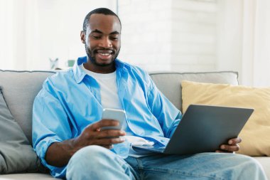 İş ve telekomünikasyon. Akıllı telefon kullanan gülümseyen siyah adam dizüstü bilgisayarlı koltukta oturuyor ve mesaj okuyor, evde çalışıyor, boş yer arıyor.