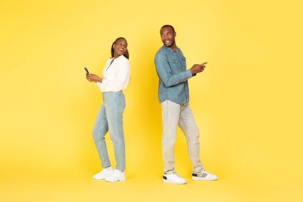 携帯電話を使用して興奮した黒のカップルは 黄色の背景の上に一緒にポーズに戻って立ちお互いに笑顔 技術とガジェット スタジオショット — ストック写真