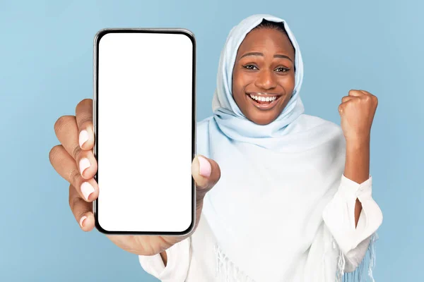 兴奋的黑人穆斯林女性展示智能手机与白色屏幕和动摇的拳头 享受销售或很酷的促销背景蓝色工作室 巨大的移动报价和应用程序广告 — 图库照片