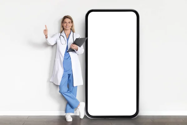 Medical App. Smiling Doctor Lady In Uniform Posing Near Big Blank Smartphone And Gesturing Thumb Up At Camera, Beautiful Female Therapist Recommending New App For Telemedicine, Collage, Mockup