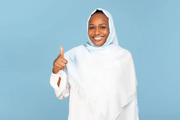青いスタジオの背景に親指を立ててカメラを見て微笑む幸せなアフリカ系アメリカ人ムスリム女性 ヒジャーブ州の陽気な黒人女性は サービスまたは購入をお勧めします — ストック写真