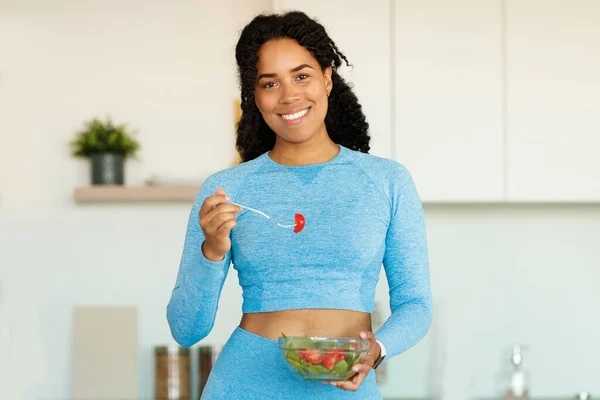 Sport and diet concept. Happy sporty black woman eating fresh salad, standing in kitchen and smiling at camera, copy space. Healthy lifestyle concept