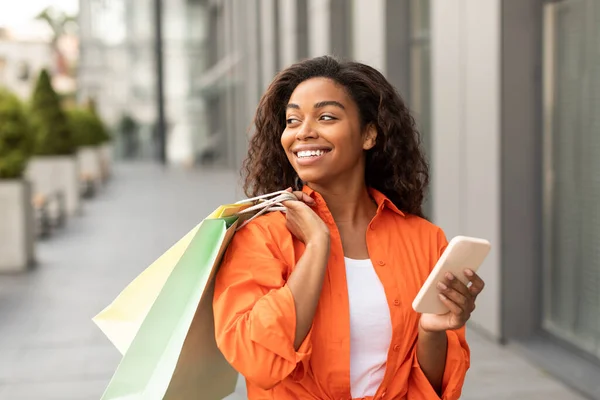 オンラインショッピング 大規模な販売 多くのショッピングバッグやスマートフォンを持ち 街の通りの空のスペースを見て カジュアルで幸せな100年アフリカ系アメリカ人女性 黒金曜日のための店からの広告とオファー — ストック写真
