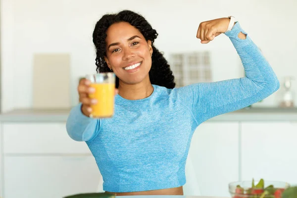 Young, strong athletic black woman in sportswear holding glass with juice and showing arm muscles, biceps, enjoying homemade beverage, standing in kitchen. Training concept