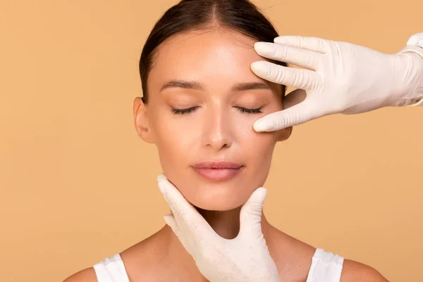 面部治疗 美容师触摸美丽的高加索女人的脸 女士紧闭双眼坐在米色背景之上 整形外科概念 — 图库照片