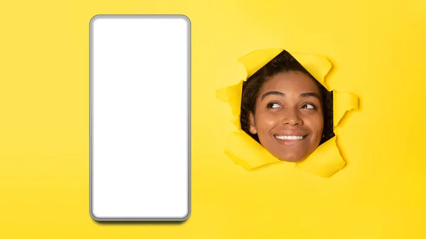 アフリカ系アメリカ人の女性は 黄色の背景にトーンペーパー広告モバイルアプリケーションで穴を通って探している空白の画面を持つ電話で見て パノラマ モックアップ — ストック写真