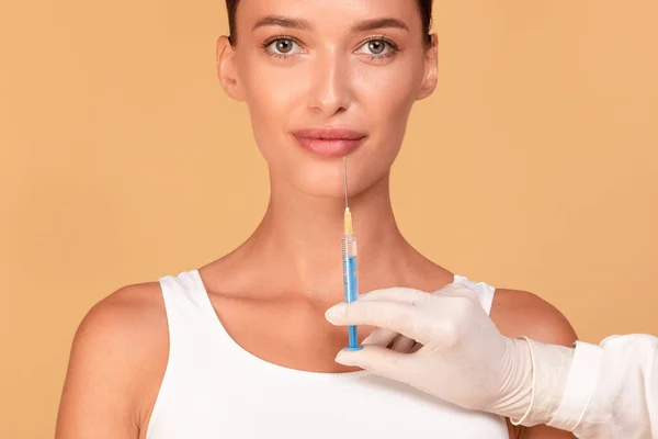 美しい若い女性は ベージュの背景に顔の注射を取得します 顎の近くに注射器を保持美容師ながら カメラを見て 化粧品手術コンセプト — ストック写真