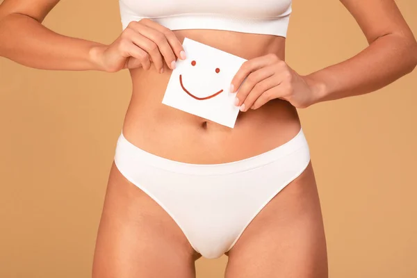 stock image Women health concept. Lady in underwear holding paper with happy smiley face in hands near abdomen zone, caucasian woman with slim body posing over beige background, cropped