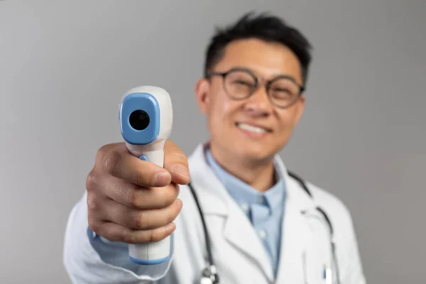 白のコートで陽気な中年中国の男性医師は 眼鏡は 灰色の背景 スタジオに隔離された非接触温度計で温度を測定します 病気や医療の治療 — ストック写真