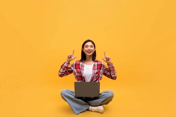 快乐的亚洲女人交叉腿坐在笔记本电脑上 指着黄色工作室背景上的空旷空间 快乐的韩国女人 有Pc广告卖座 — 图库照片