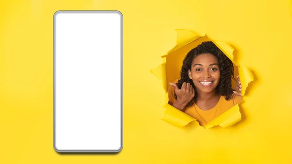 肯定的な黒の女性は 空白の画面でスマートフォンを表示します 黄色い背景の上に置かれたトーン紙を通して見てガジェットで指を指す スタジオショット このモバイルオファーを見てください パノラマ — ストック写真
