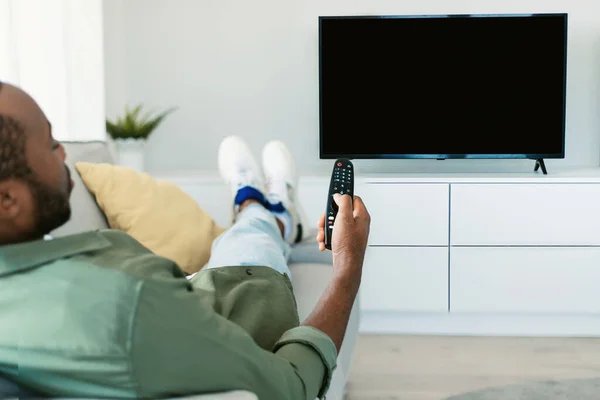 Back view of black man watching TV pointing remote control at empty plasma screen, switching channels, resting on sofa at home. Guy enjoying movie or sport program, using television set. Mockup