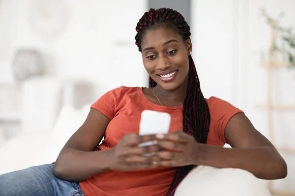 年轻美丽的黑人女人在家里放松的时候用智能手机发短信 非洲裔美国女人坐在沙发上微笑 用手机上网聊天 复制空间 — 图库照片