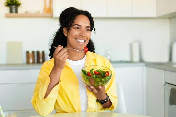 Excited black lady sitting in kitchen and eating fresh vegetable salad, looking aside and smiling, healthy woman enjoying food full of vitamins, copy space