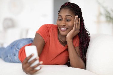 Gülümseyen siyah kadın evdeki rahat koltukta akıllı telefonuyla rahatlıyor, mutlu Afrikalı genç Amerikalı bayan modern Gadget ile koltukta dinleniyor, cep telefonuyla interneti tarıyor,