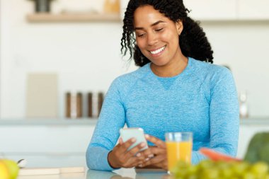 Elinde akıllı telefon tutan, elinde taze meyve suyu ve sebzelerle mutfak masasında oturan pozitif siyah kadın. Evde sağlıklı kahvaltı ederken sosyal medyayı kontrol eden kadın, boşluğu kopyala
