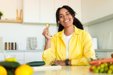 Mutlu Afro-Amerikan ev hanımı bir dilim salatalık tadıyor, taze sebze salatası hazırlıyor, sağlıklı beslenmenin tadını çıkarıyor, mutfakta iç kısımda oturuyor ve kameraya gülümsüyor.