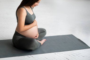 Doğum Konsepti. Yoga minderi üzerinde oturan ve göbeğini okşayan, aktif giyimli, güzel, karnı ağrıyan, gülümseyen, hamilelerin tadını çıkaran hamile bir kadın.