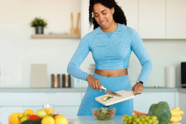 Mutfakta durmuş taze sebze salatası hazırlayan spor giysili narin Afro-Amerikan kadın antrenmandan sonra sağlıklı yemeğin tadını çıkarıyor. Fitness, diyet ve sağlıklı yaşam tarzı konsepti