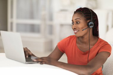 Video çağrısı. Kulaklık takmış genç siyahi bir kadın dizüstü bilgisayarında telekonferans yapıyor. Evde oturmuş, bilgisayarını online iletişim için kullanan Afrikalı Amerikalı bir kadın gülümsüyor.