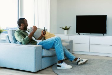Siyahi adamın TV 'de spor maçı izlerken boş ekranda zaferi kutlarken, yumruklarını sallarken, oturma odasında kanepede otururken. Model