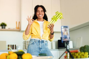 Yemek blogu. Mutfakta telefon kamerasıyla çekim yapan mutlu siyahi kadın sosyal medya için yemek pişiriyor ve video kaydediyor. Tripodla akıllı telefona nadiren Afrikalı Amerikalı kadın gösteriyor.