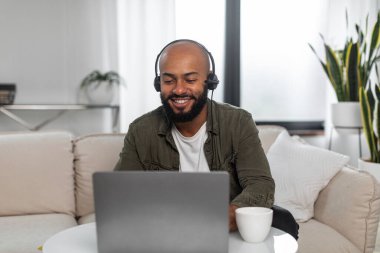 Laptopta çalışan, gülümseyen, kulaklık takan, evdeki koltukta oturan mutlu Latin adam. Başarılı bir kariyer ve mesafeli iş kavramı