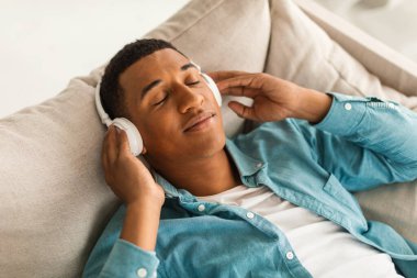 Kot pantolonlu, kablosuz kulaklıklı, koltukta uzanan, favori müzikten hoşlanan, rahat ve tembel bir erkek. Ses uygulaması ile evde dinlen ve rahatla