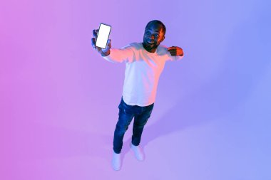 Neşeli, çekici, şık, Afro-Amerikalı olgun bir adam beyaz ekranlı cep telefonunu gösteriyor, iyi bir teklif veya mobil uygulama öneriyor, model, neon stüdyo arka planı, yüksek açılı çekim, kopyalama alanı