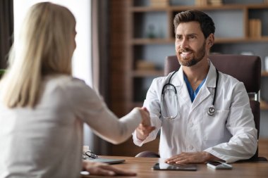 Ofiste buluşurken üniformalı, kadın hastayla tokalaşan, beyaz önlüklü ve steteskoplu gülümseyen, klinikte tanınmayan bir kadını selamlayan dost canlısı erkek doktor.