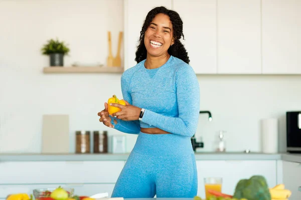 Excited sporty black lady cooking fresh vegetables salad and laughing at camera, standing in kitchen, preparing healthy lunch. Fit woman having healthy meal after workout