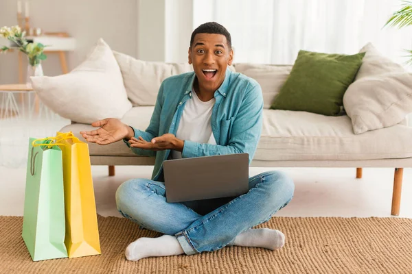 ラップトップ付きの喜んで興奮した若い黒人男性は リビングルームのインテリアで床に座って 購入とパッケージの多くを示しています オンラインショッピングからの感情 お店のための巨大な販売 納期Covid — ストック写真