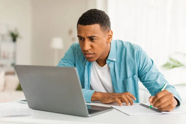 Serious concentrated bored young african american male in jeans looking at laptop making notes, find mistake in room office interior. Problems at work, business, freelance and study at home covid-19