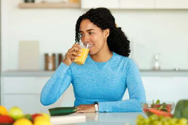 Detox concept. Excited black lady drinking tasty homemade juice, sitting at kitchen table, preparing lunch after domestic workout, free space