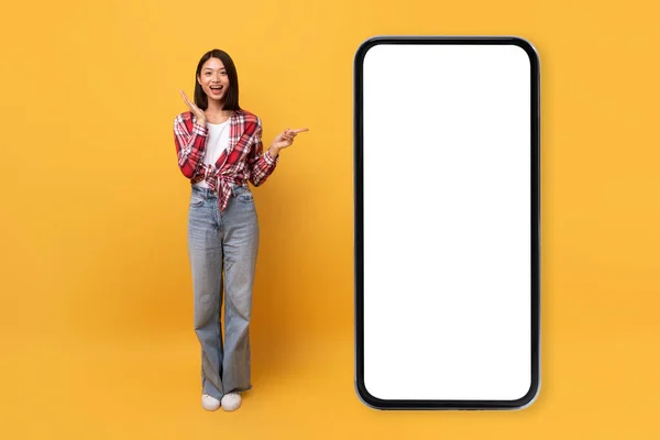 移动报价 兴奋的亚洲女人一边指着空白屏幕的大型智能手机 一边推荐新的应用程序或网站 站在黄色的背景上 全身而退 莫科普 — 图库照片