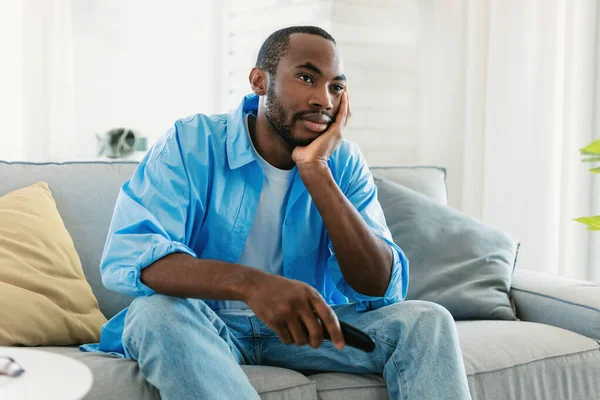 テレビ番組の放送を中止 テレビを見ている不満を持つ若い黒人男性 リモコンでチャンネルを切り替える 自宅でソファに座って 孤独を感じます フリーコピースペース — ストック写真
