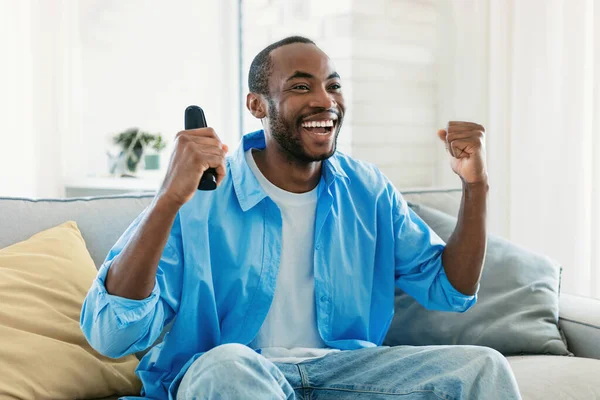 喜形于色的黑人男子一边看电视 一边高兴地挥动拳头 拿着遥控器 坐在客厅的沙发上 电视节目 胜利庆祝概念 — 图库照片