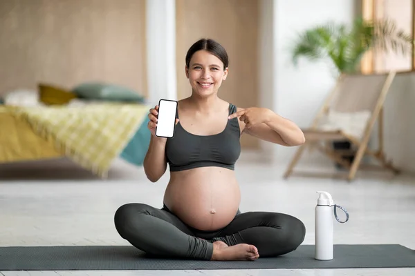 フィットネスアプリ 笑顔妊娠中の女性はヨガマットに座って 白い画面で空白のスマートフォンを表示 モバイルアプリケーションやウェブサイトのデザインのためのコピースペースを実証幸せな期待の女性 モックアップ — ストック写真