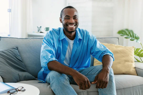 Portrait of handsome black man smiling and looking at camera, sitting on sofa at home, free space. Happy african american guy expressing positive emotion, spending time indoors