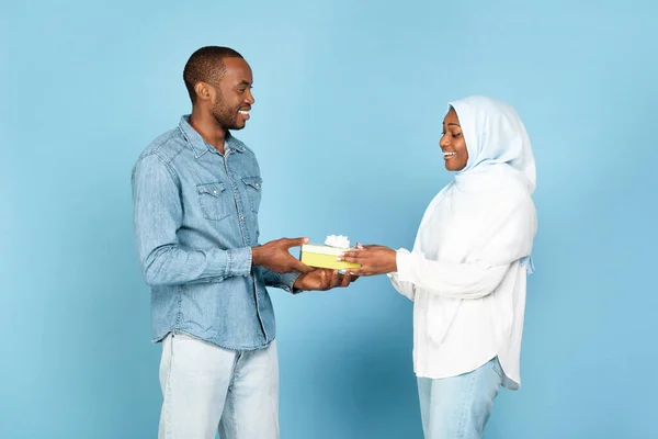 Cheerful Black Muslim Husband Giving Present Congratulating His Wife On Birthday, Celebrating Holiday Together Standing Over Blue Studio Background. Gifts Delivery Concept