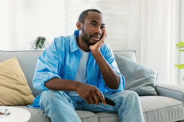 一个无聊的非洲裔美国人在家里看电视 坐在沙发上 头靠在手上 自由自在的形象 带遥控器的不满意的黑人交换信道 — 图库照片