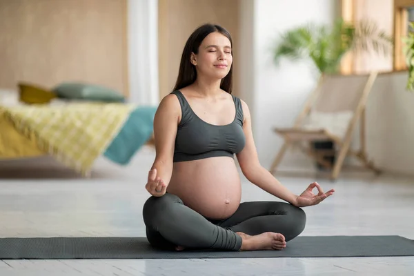 漂亮的年轻孕妇在家里练习瑜伽 在客厅的健身垫上坐着荷花 期待着女性闭眼沉思 沉思着健康 — 图库照片
